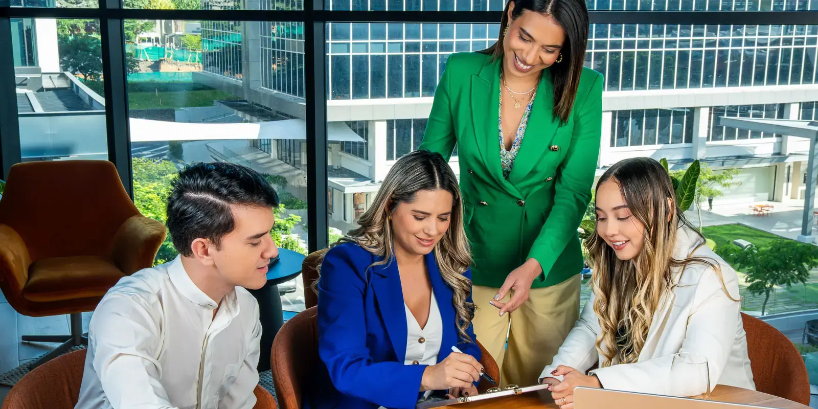 Grupo de cuatro personas reunidas en una mesa de oficina, colaborando y revisando documentos frente a una ventana con vista a edificios.