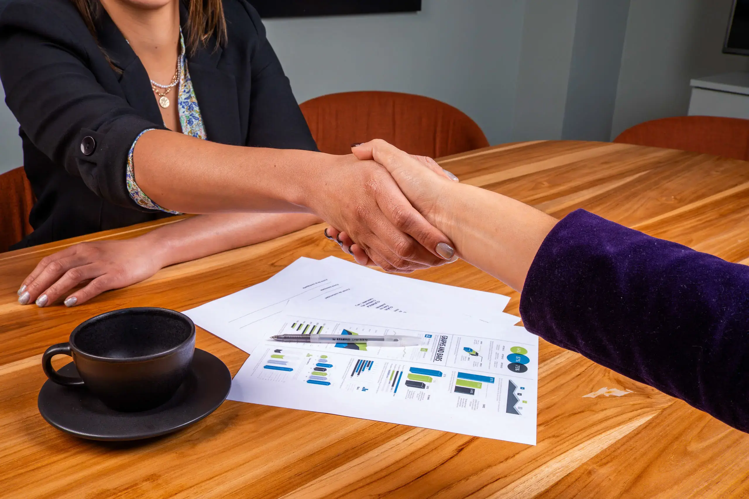 Dos personas dándose la mano sobre una mesa con documentos y una taza negra.
