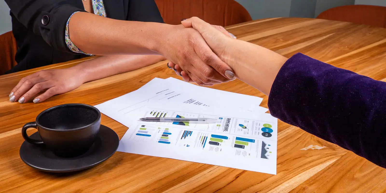 Dos personas dándose la mano sobre una mesa con documentos y una taza negra.