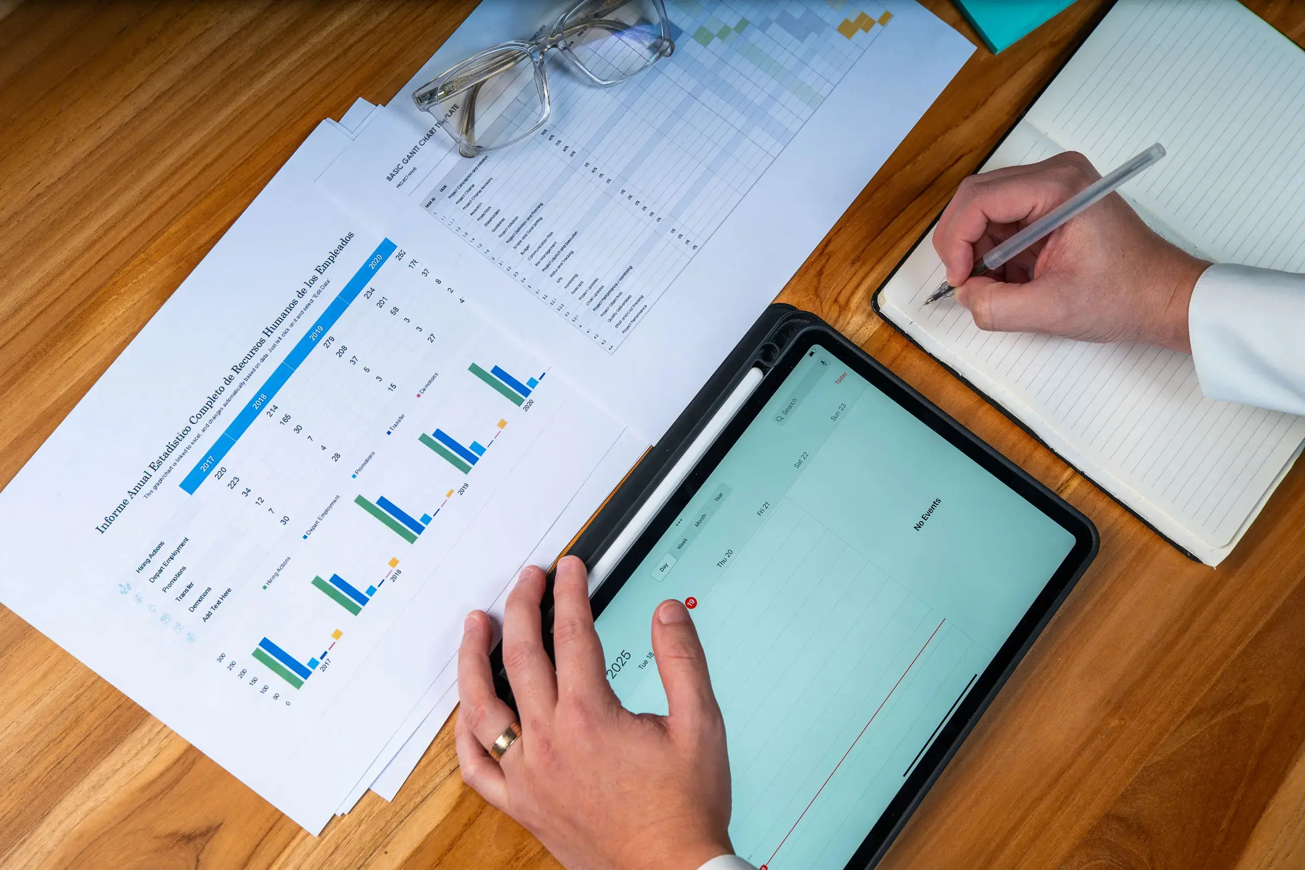 Manos trabajando con una tablet, documentos con gráficas y un cuaderno sobre una mesa de madera.