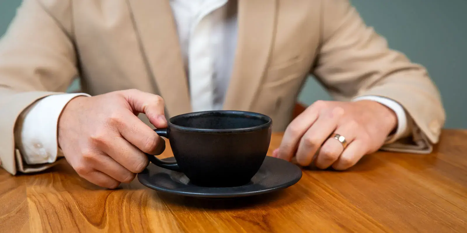 Persona con saco beige sosteniendo una taza negra sobre una mesa de madera.