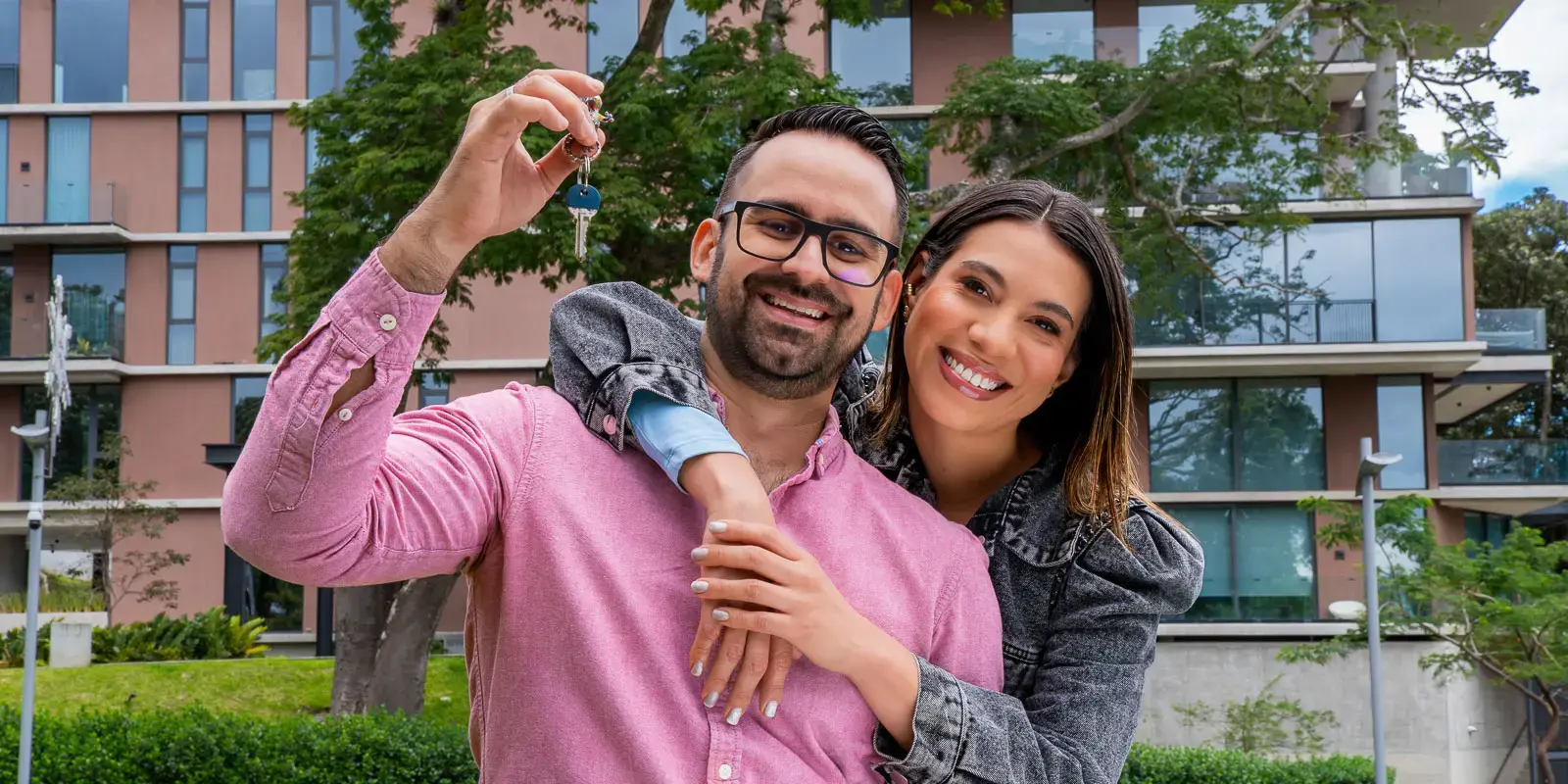 Pareja sonriendo mientras el hombre sostiene un manojo de llaves frente a un edificio residencial.