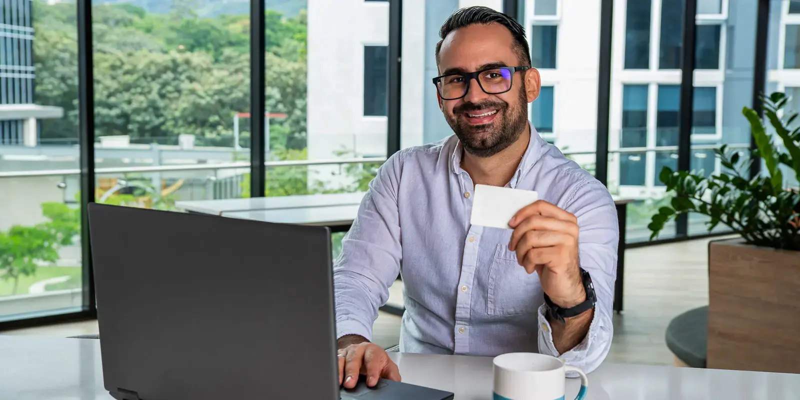Hombre sentado frente a una laptop sosteniendo una tarjeta en la mano en un entorno de oficina con ventanales.