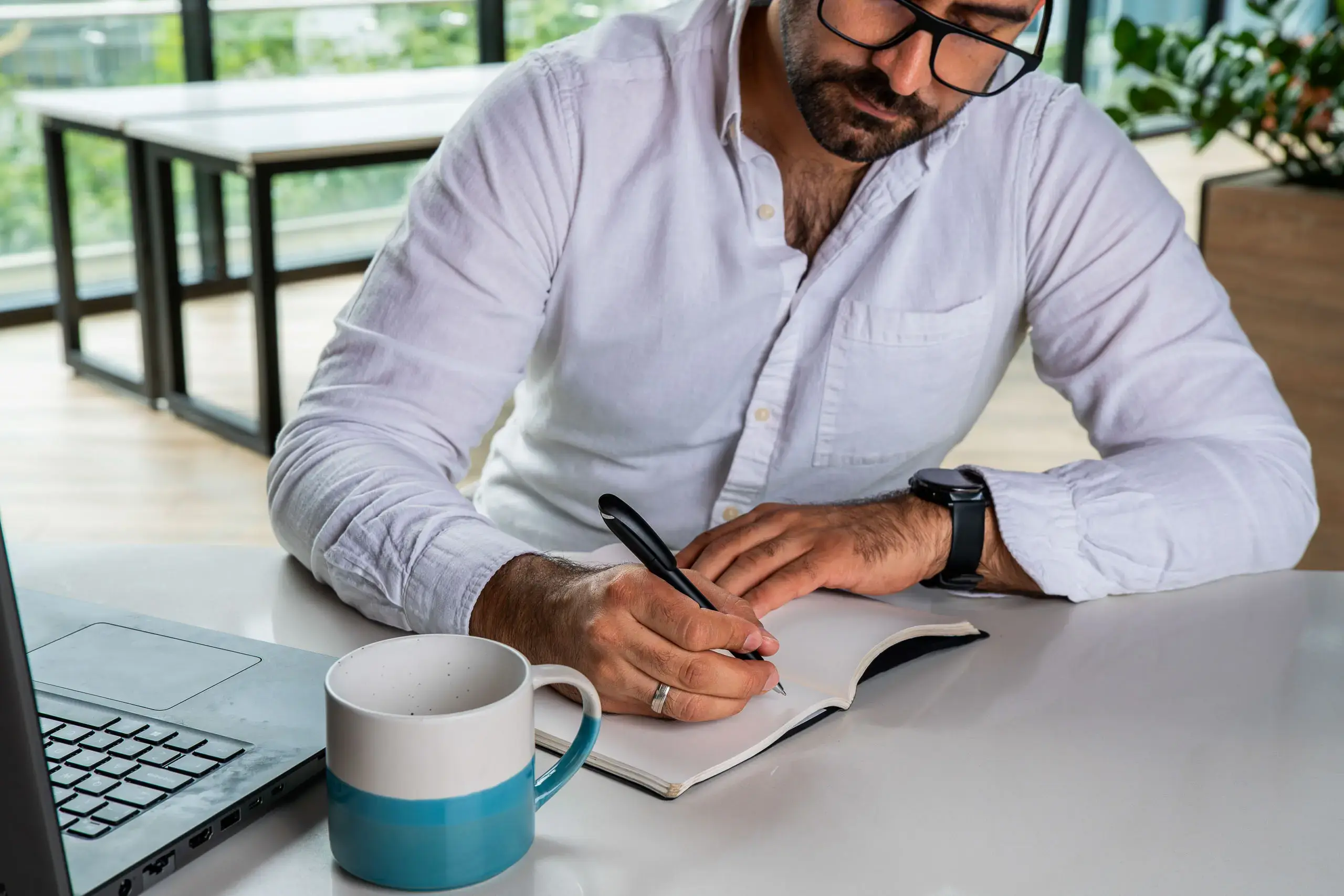 Hombre escribiendo en una libreta sobre un escritorio junto a una computadora portátil y una taza.