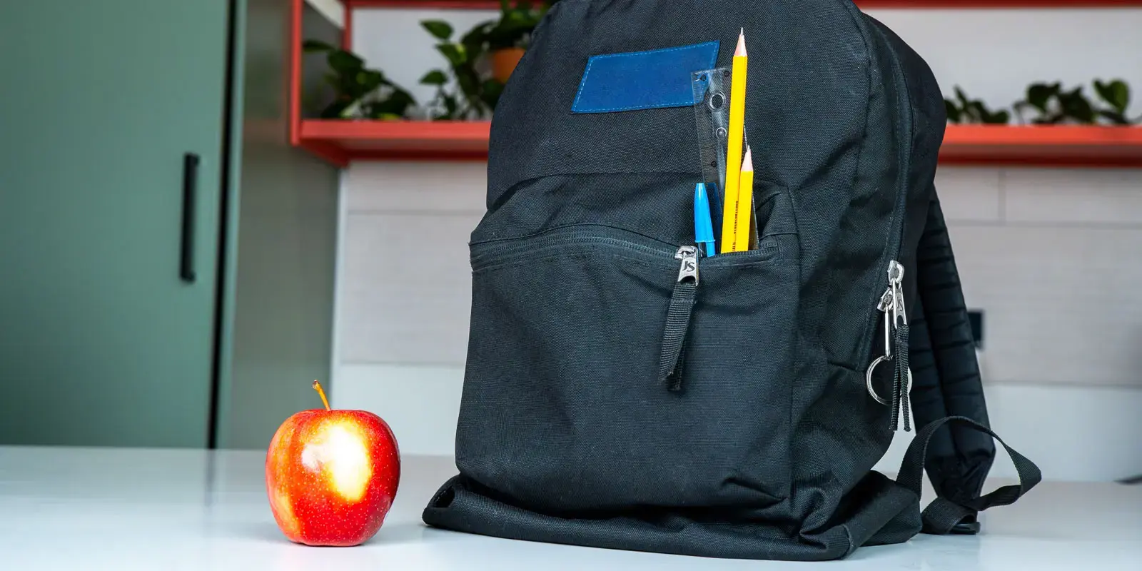 Mochila negra con lápices y bolígrafos en el bolsillo frontal junto a una manzana roja sobre una mesa.