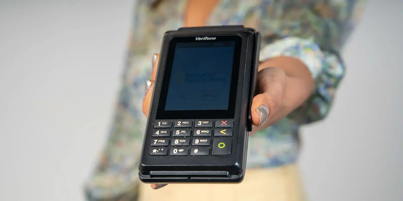 Terminal de pago Verifone sostenido al frente por una persona, con pantalla mostrando el logo del Banco Nacional.