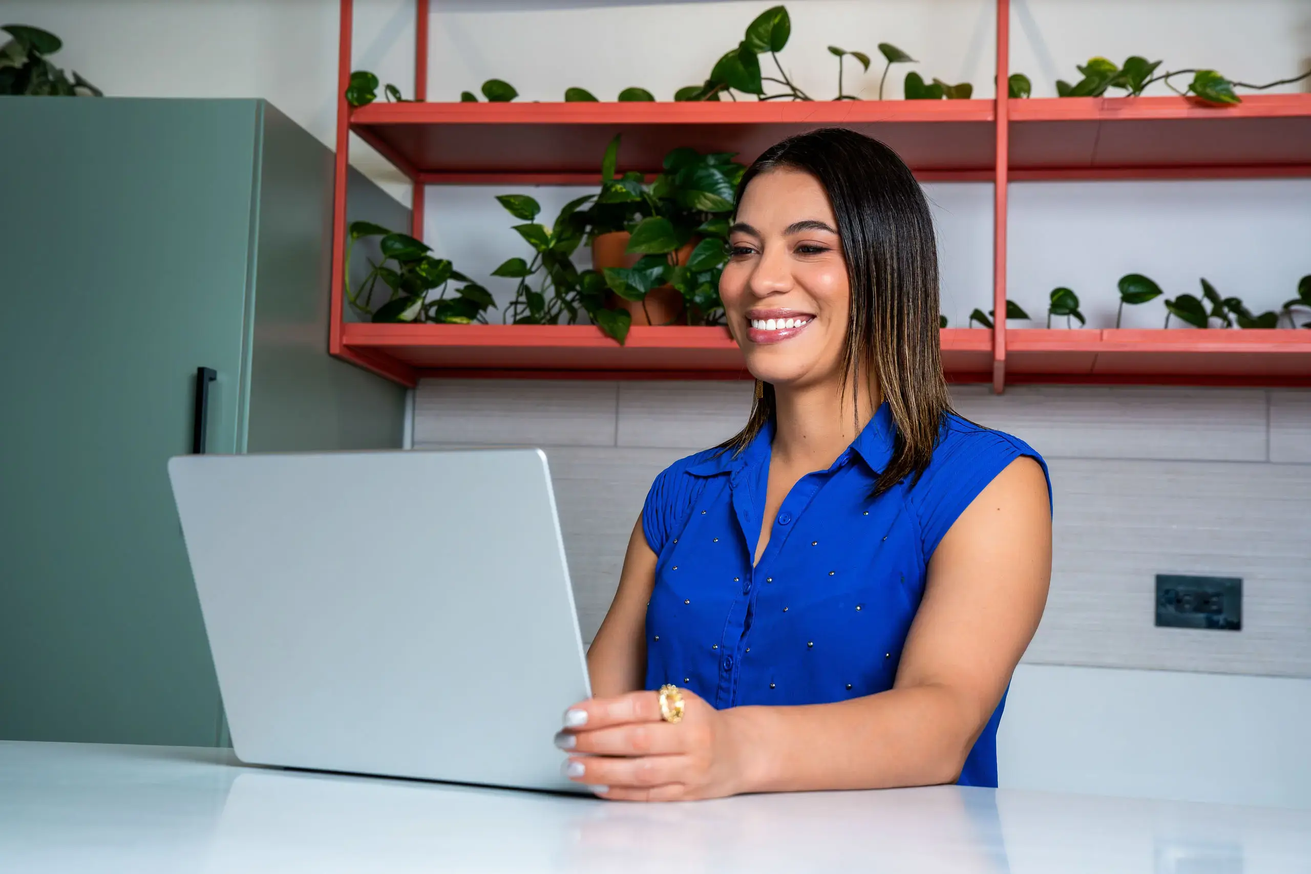 Mujer sonriendo mientras trabaja en una laptop en una oficina con estantes y plantas.