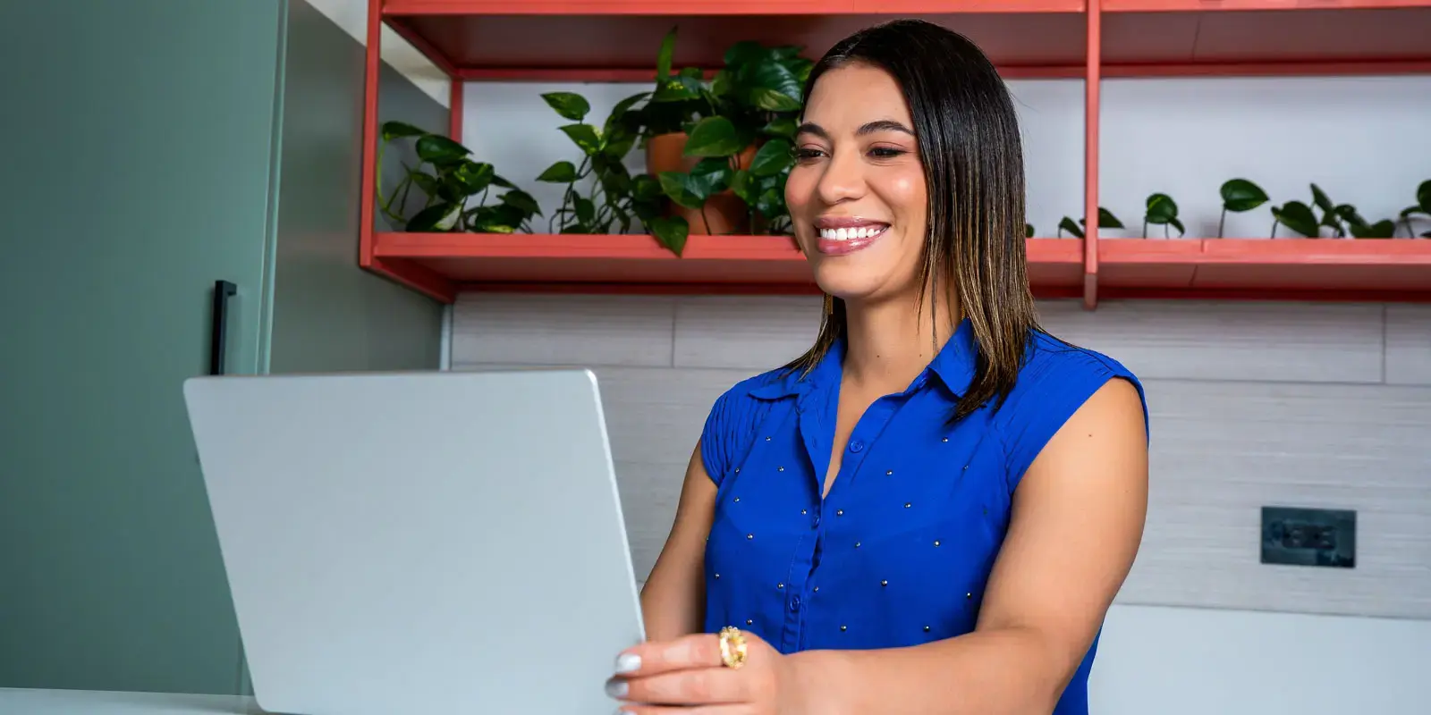 Mujer sonriendo mientras trabaja en una laptop en una oficina con estantes y plantas.