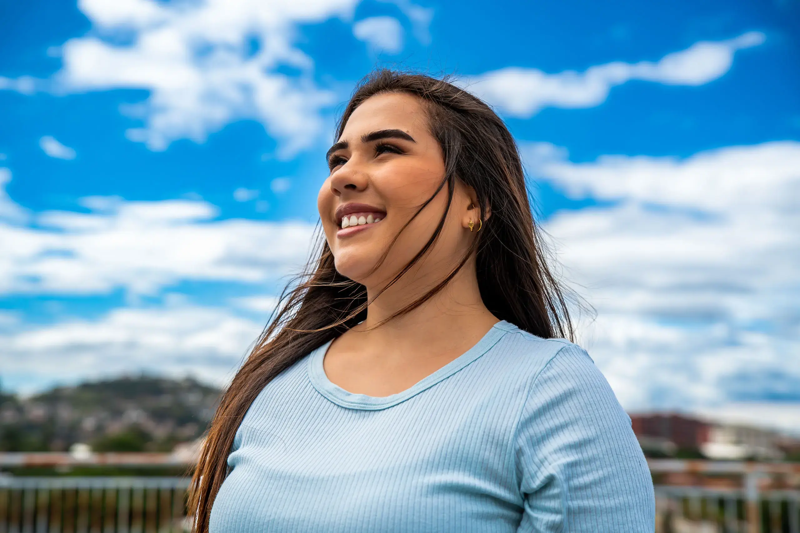 Mujer sonriendo al aire libre con el cielo azul y nubes de fondo.