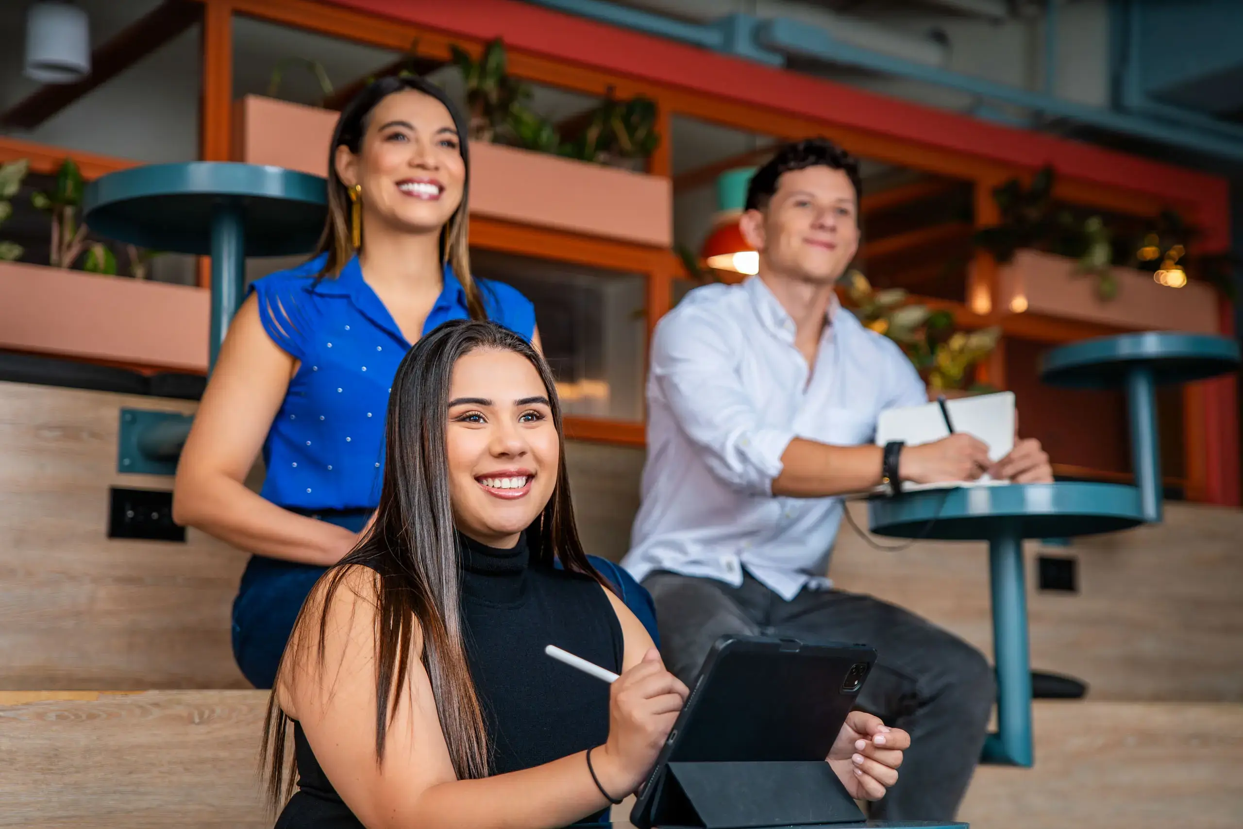 Tres personas sentadas en un espacio interior, sonriendo mientras toman notas y usan una tablet.