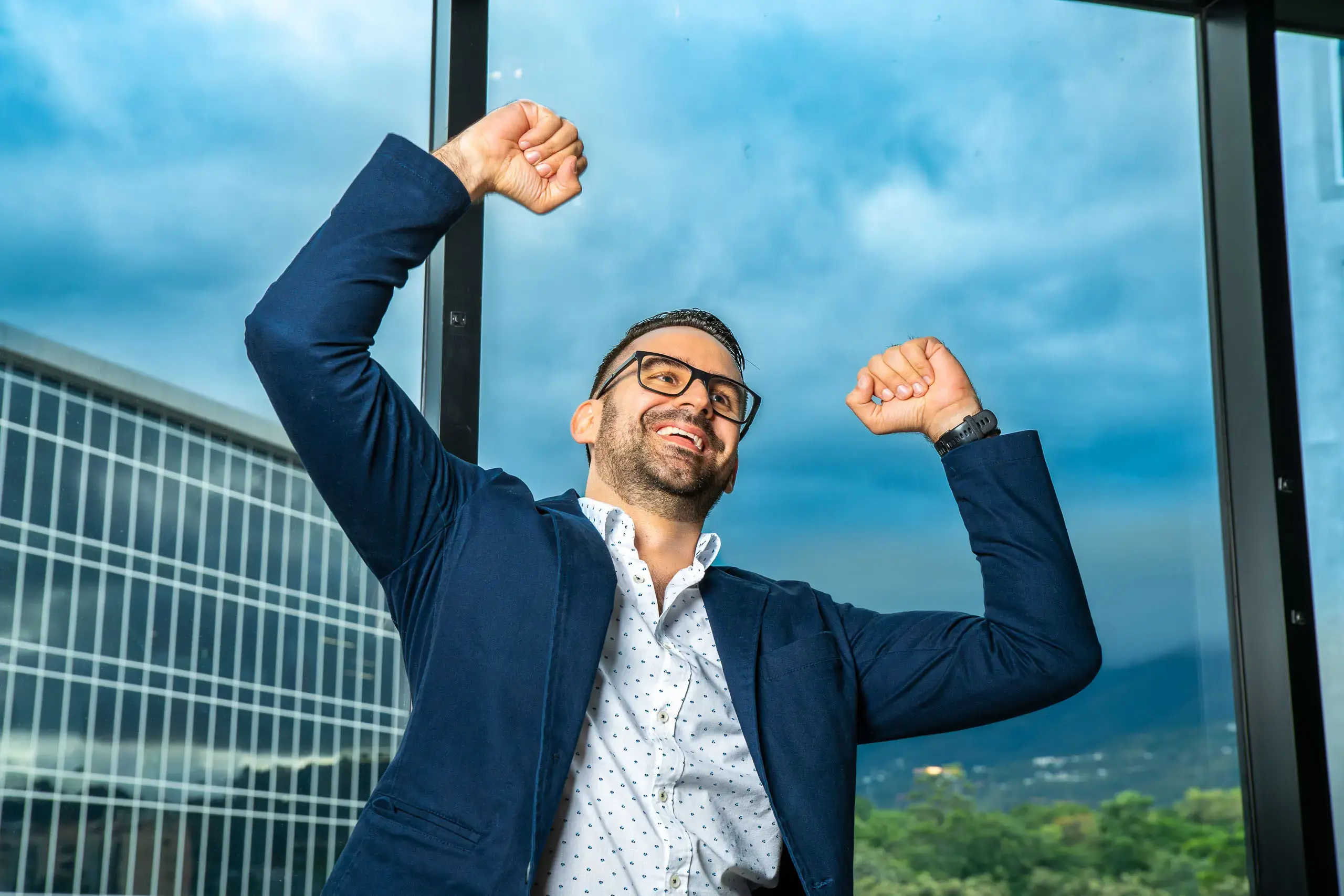 Hombre levantando los brazos con expresión de alegría frente a una ventana con vista a edificios y montañas.