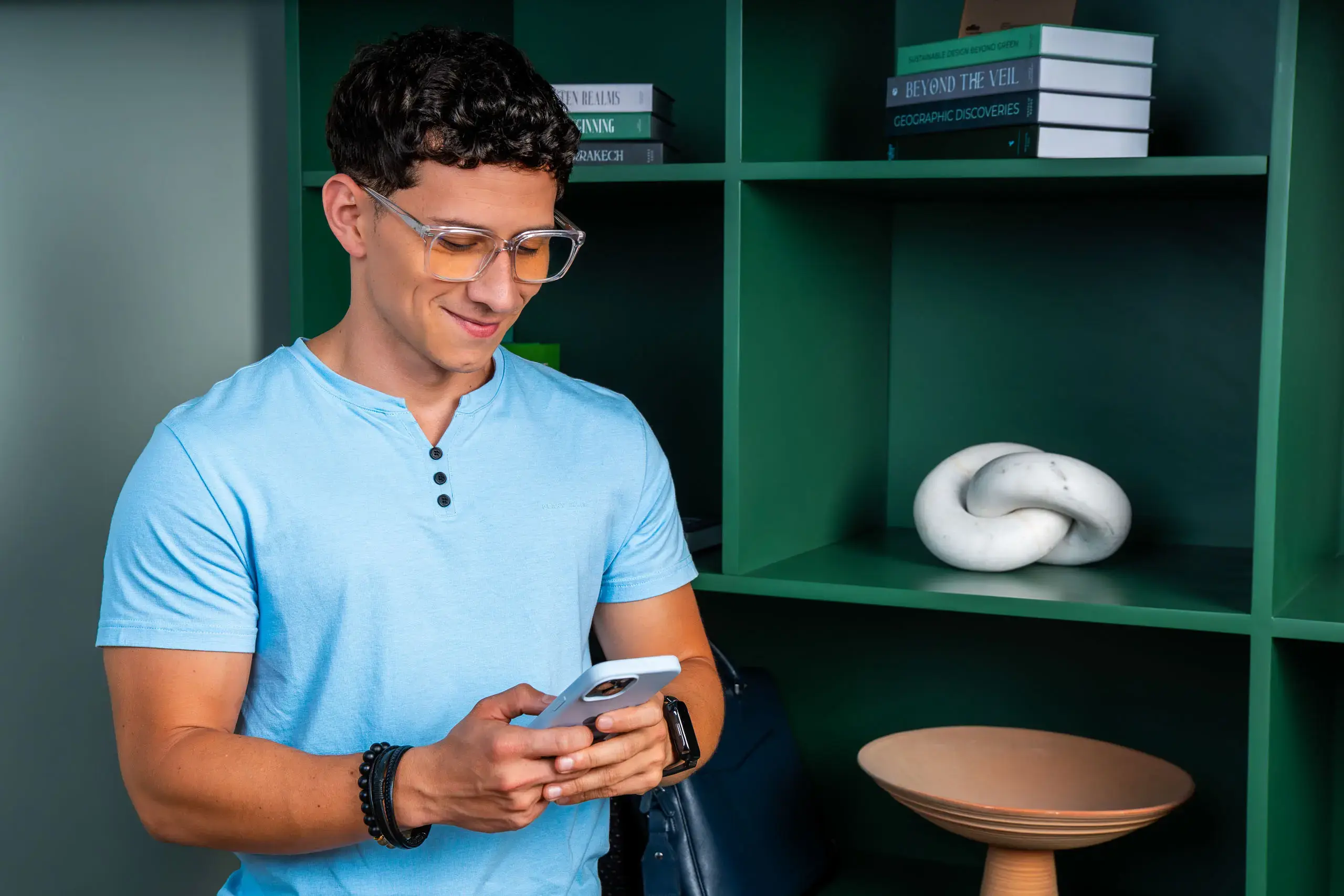 Hombre joven con gafas usando un teléfono móvil frente a un estante verde.