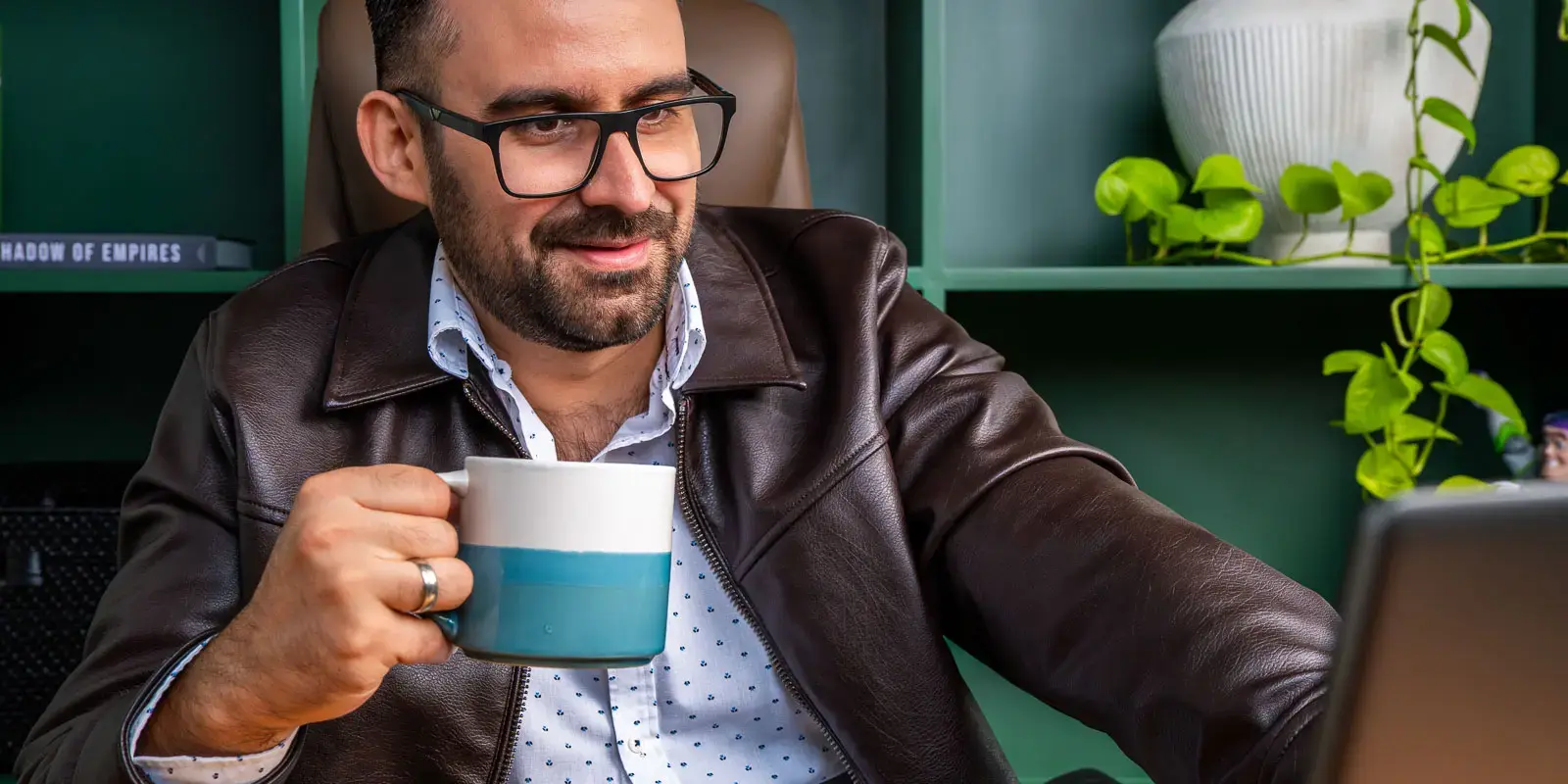 Hombre con gafas y chaqueta de cuero sosteniendo una taza mientras trabaja en una computadora portátil.