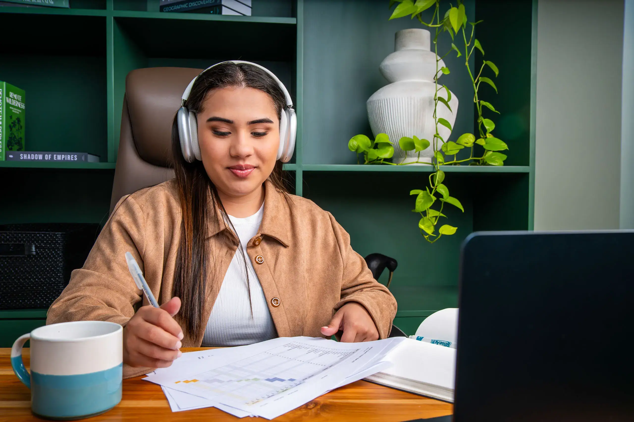 Mujer con audífonos revisando documentos y tomando notas frente a una computadora en una oficina.