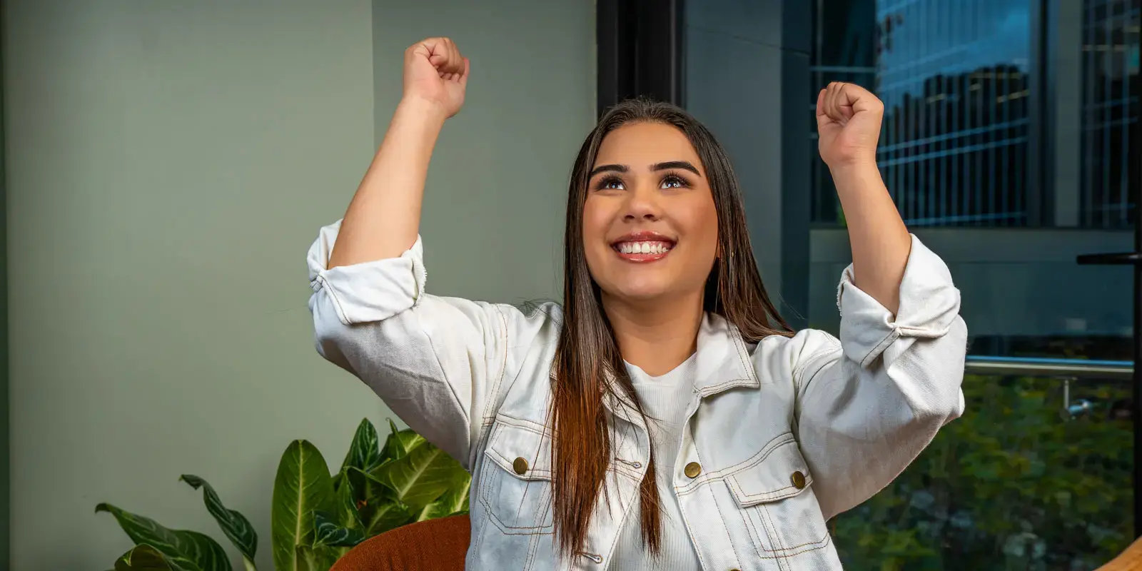 Mujer joven levantando los brazos con expresión de alegría en un espacio interior con ventanales.