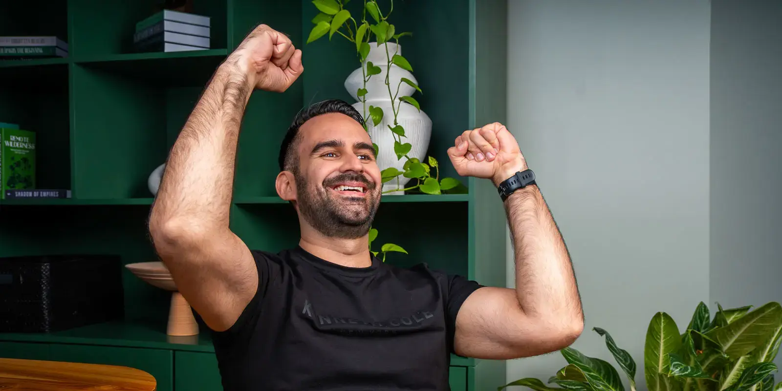 Hombre levantando los brazos con gesto de alegría en un espacio interior con estantería verde y plantas.