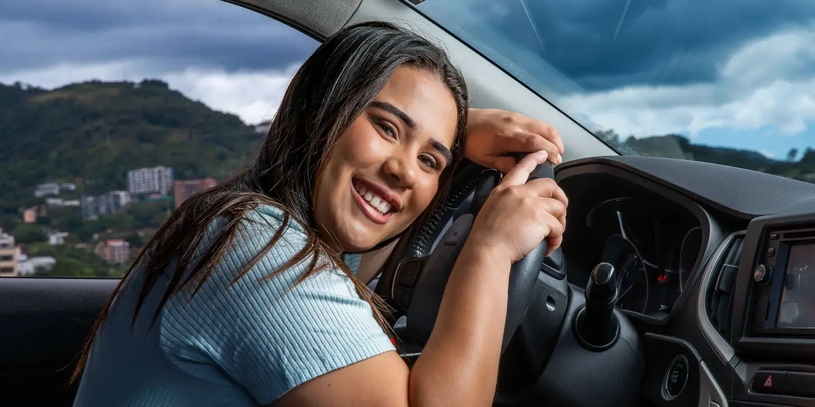 Mujer sonriendo mientras sostiene el volante dentro de un automóvil con paisaje urbano al fondo.