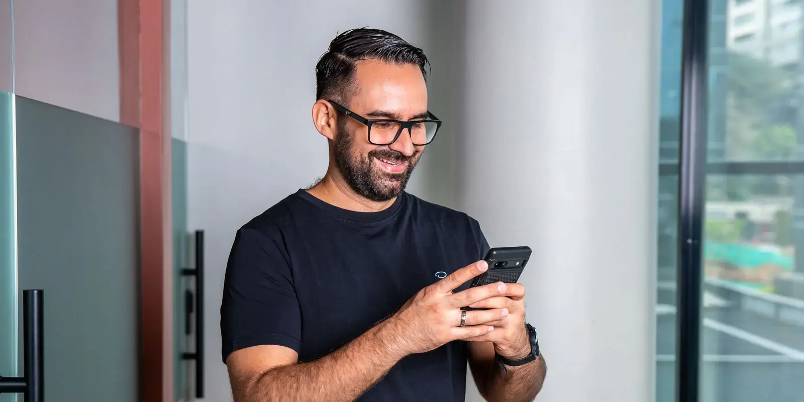 Hombre sonriendo mientras usa su teléfono móvil en un entorno de oficina con ventanales.