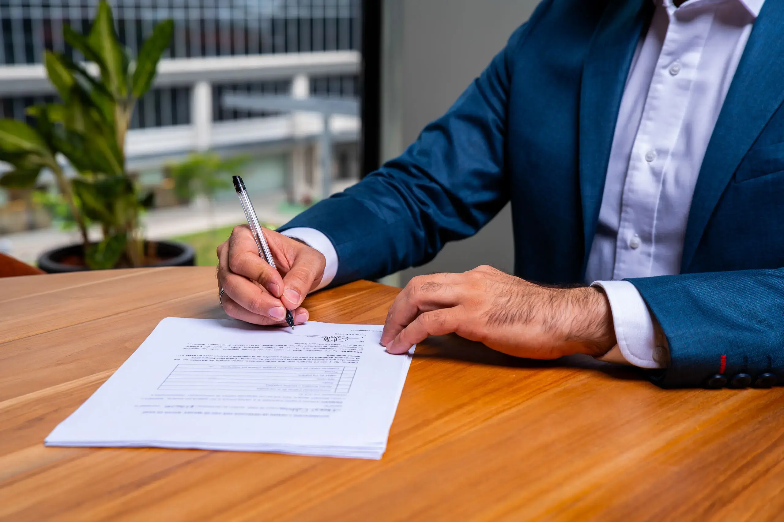 Manos de un hombre firmando un documento sobre una mesa de madera en una oficina.