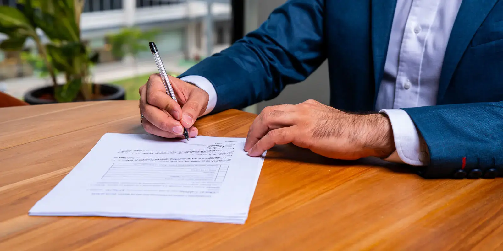 Persona firmando un documento sobre una mesa de madera, vistiendo un traje azul.