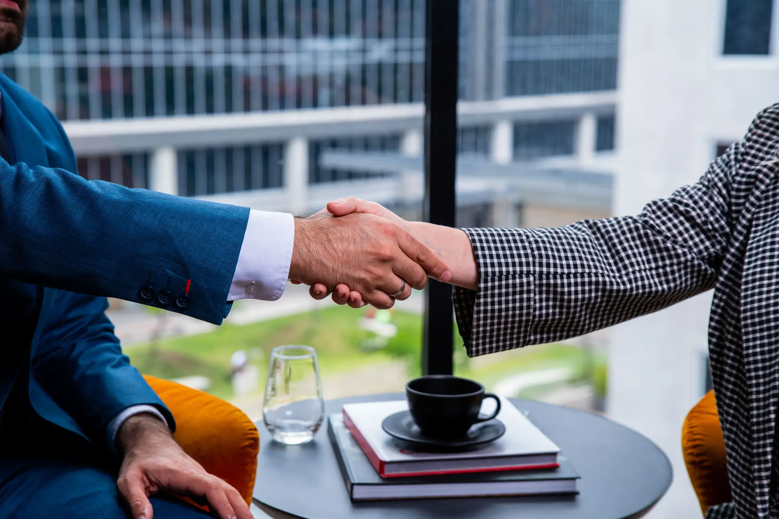 Dos personas estrechando las manos sobre una mesa con una taza y libros en un espacio con ventanales.