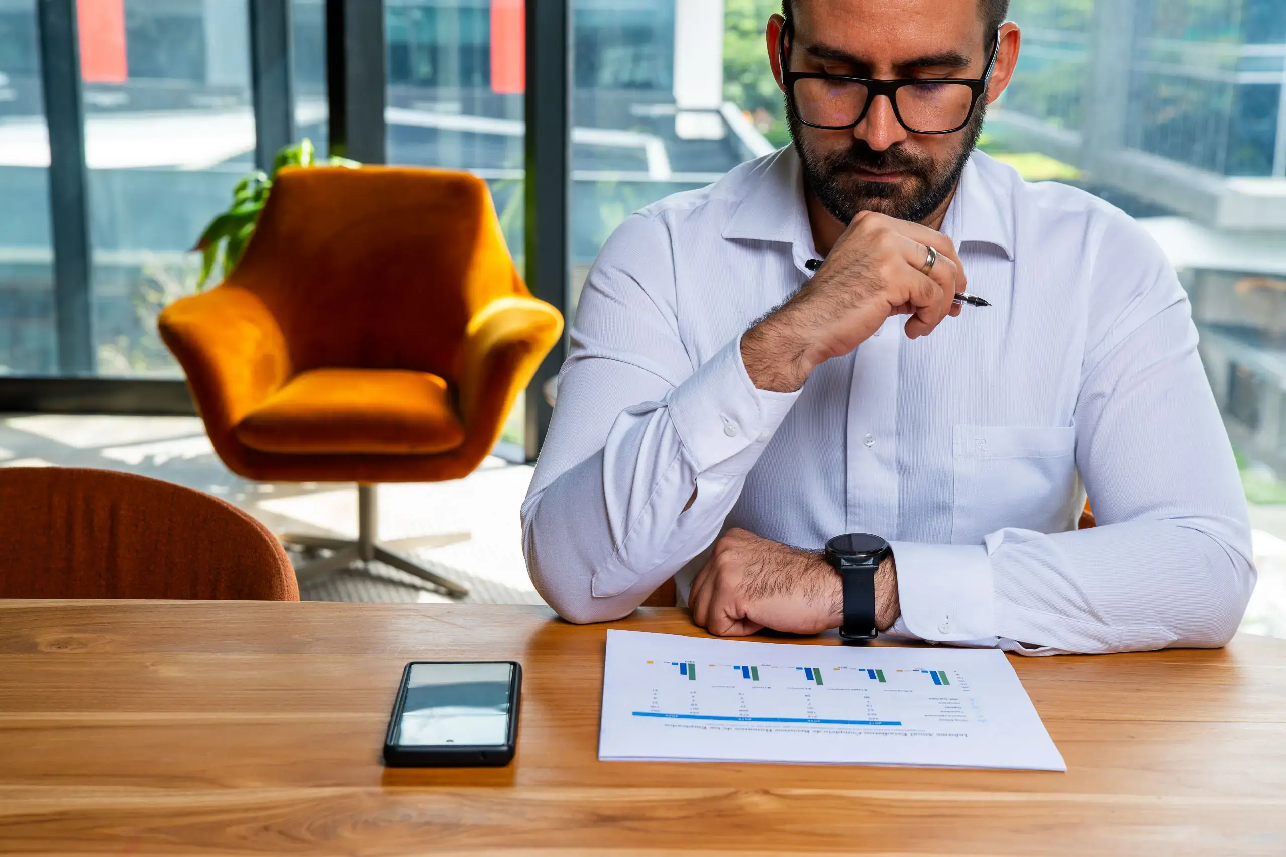 Hombre con camisa blanca revisando un documento con gráficas en una oficina, con un teléfono móvil sobre la mesa.