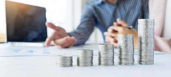 Montones de monedas apiladas en crecimiento progresivo sobre una mesa, con una persona desenfocada al fondo gesticulando, simbolizando incremento financiero.