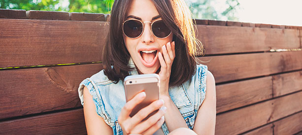 Joven sorprendida mirando su celular mientras está sentada frente a una cerca de madera.