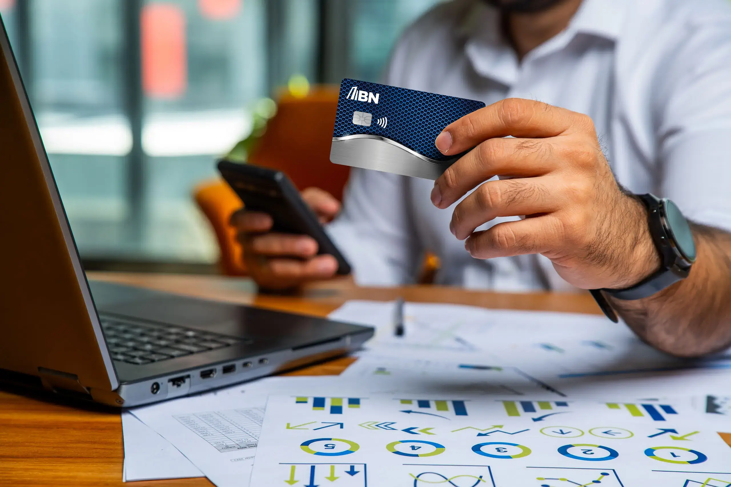 Hombre sosteniendo una tarjeta BN Prepago mientras usa su teléfono frente a una laptop y documentos con gráficas.