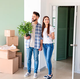 Pareja joven entrando a su nuevo hogar con cajas de mudanza y una planta en mano.