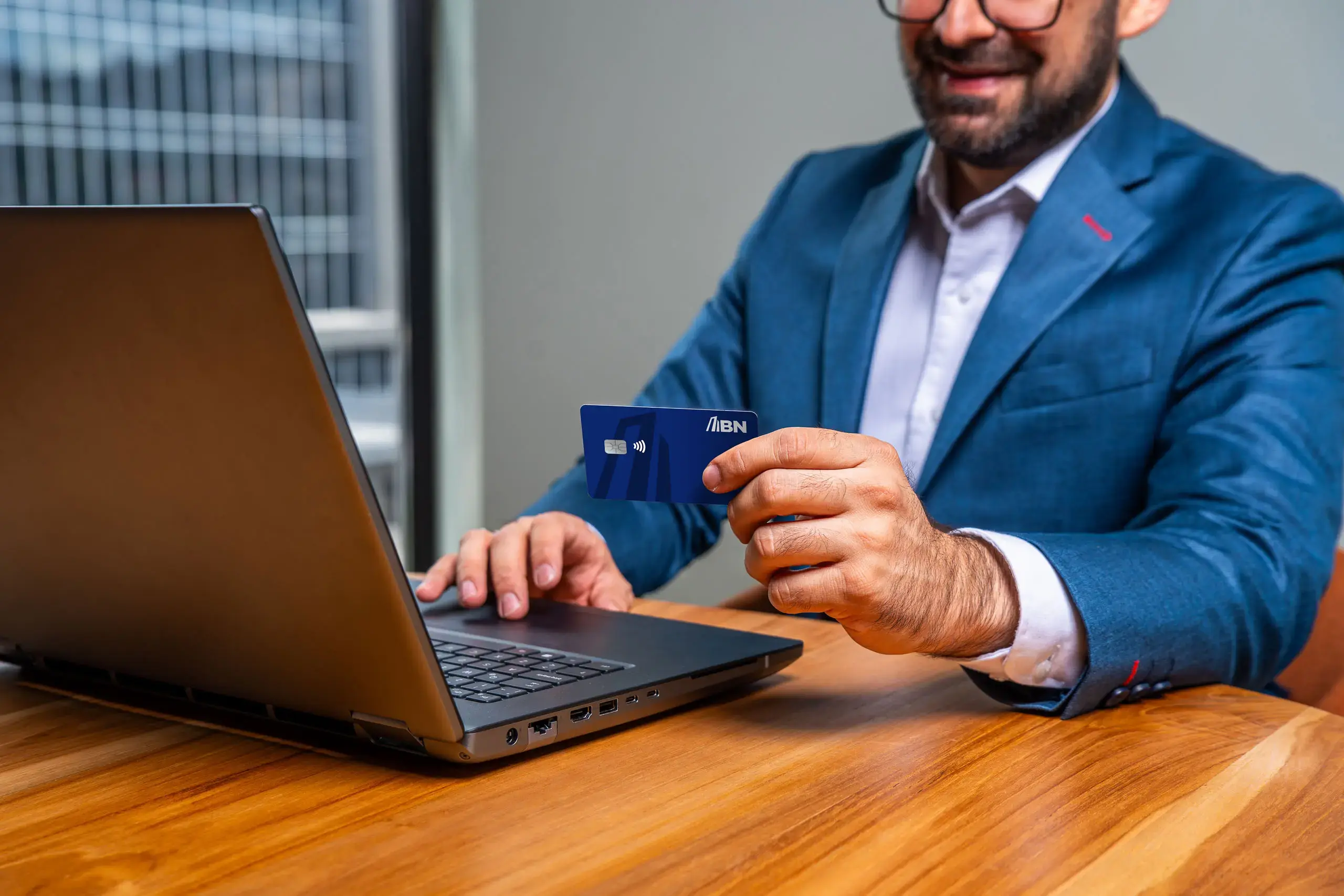 Hombre con traje azul sosteniendo una tarjeta BN azul frente a una computadora portátil sobre una mesa de madera.