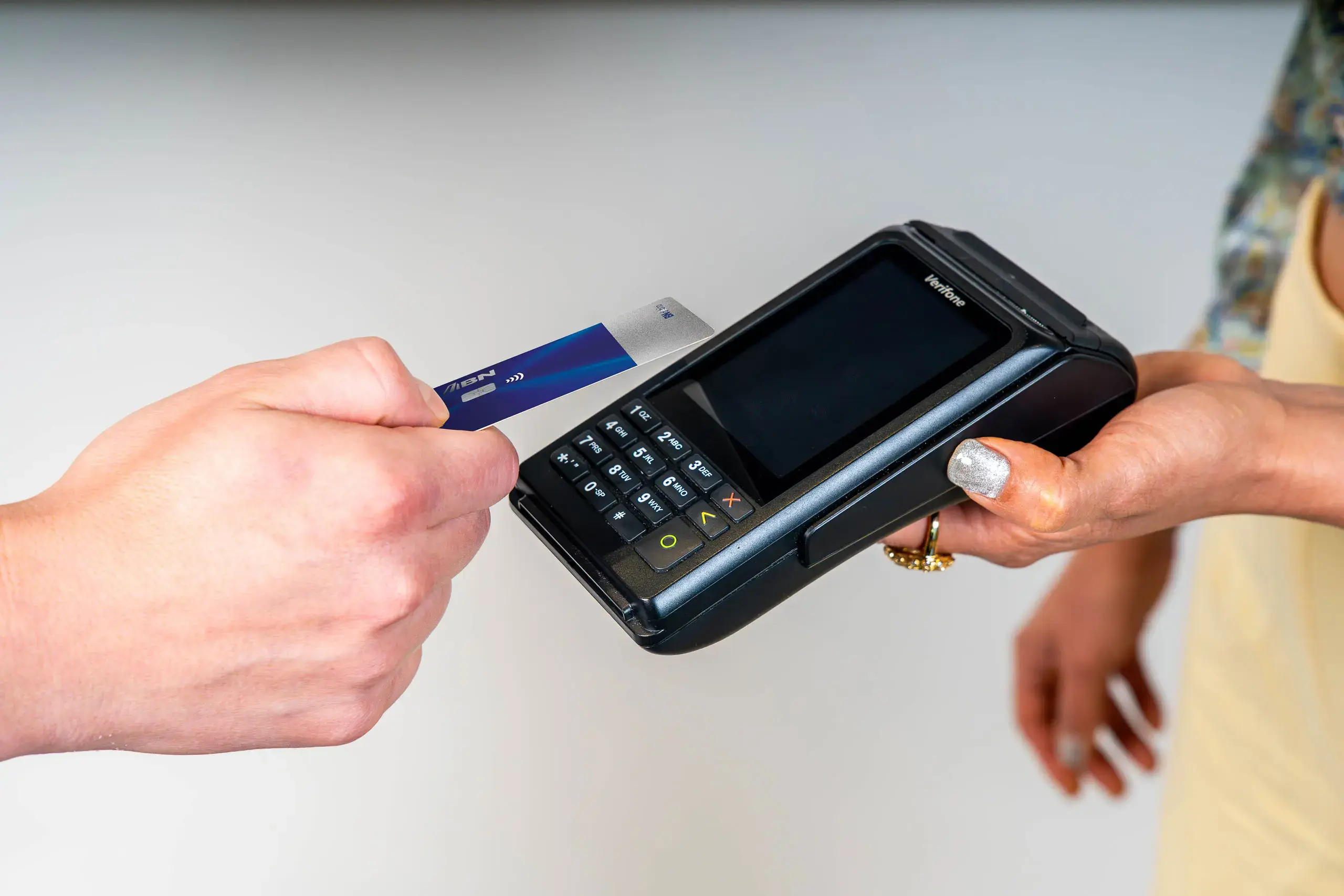 Mano acercando una tarjeta azul BN Visa a un datáfono Verifone sostenido por otra persona.