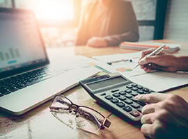 Persona usando calculadora y escribiendo en documentos financieros junto a una laptop y lentes sobre el escritorio.