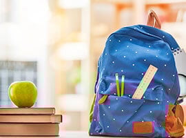 Mochila escolar azul con útiles escolares, colocada junto a una manzana verde y libros.