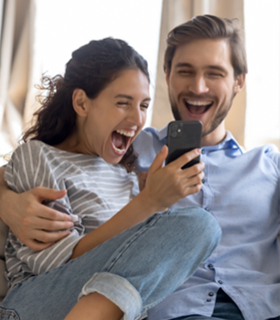 Una pareja sonriente y emocionada observa la pantalla de un teléfono móvil, mostrando entusiasmo por una buena noticia digital.