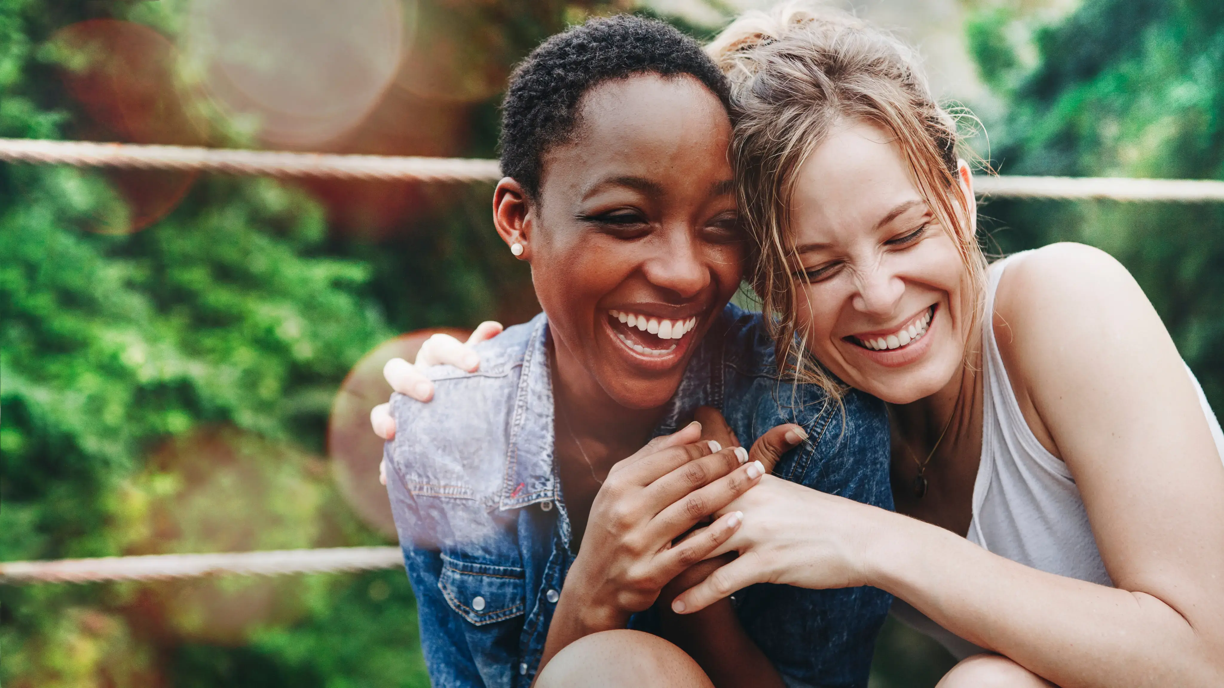 Dos amigas abrazadas y riendo al aire libre frente a un fondo verde.