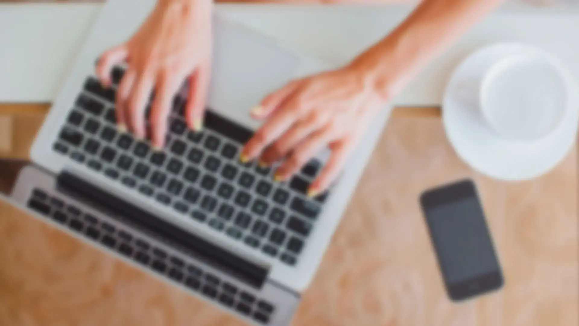 Persona escribiendo en una laptop sobre un escritorio con un celular y una taza al lado, vista superior desenfocada.