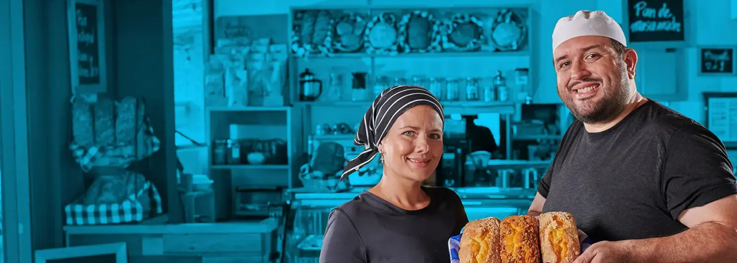 Panaderos sonrientes sosteniendo una bandeja con panes artesanales, de pie frente al mostrador de una panadería con estanterías llenas de productos al fondo.
