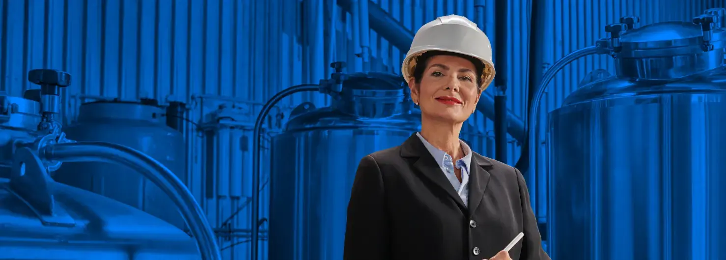 Mujer ejecutiva con casco de seguridad en una planta industrial, de pie frente a tanques de acero inoxidable sobre un fondo azul.