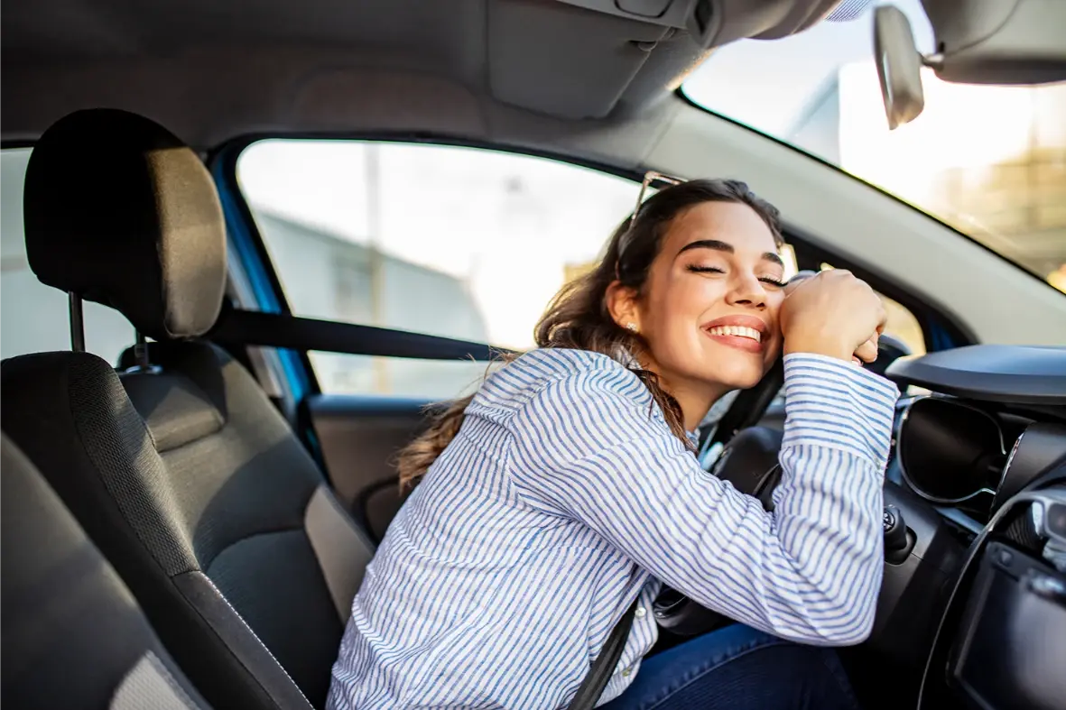 Mujer joven sonriendo mientras abraza el volante dentro de un automóvil.