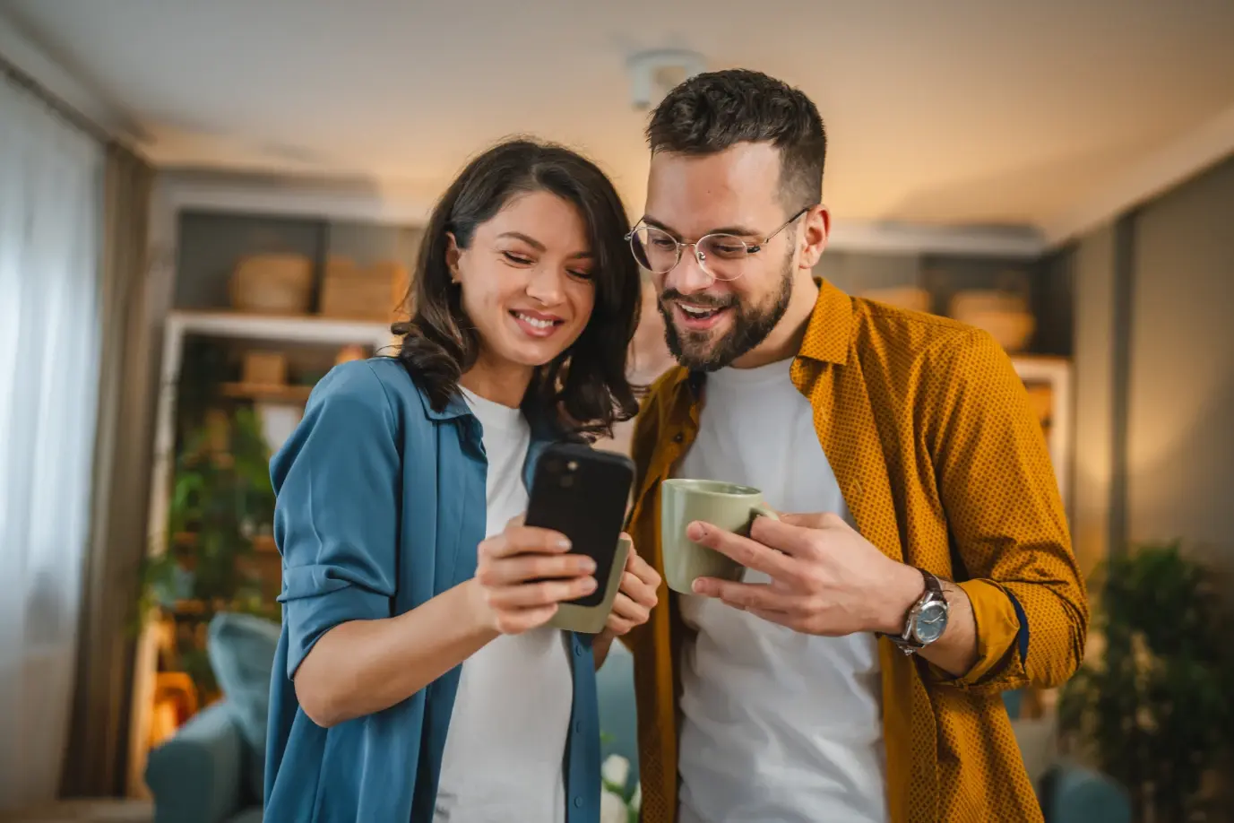 Pareja observando la pantalla de un teléfono móvil mientras sostienen tazas dentro de una sala de estar.