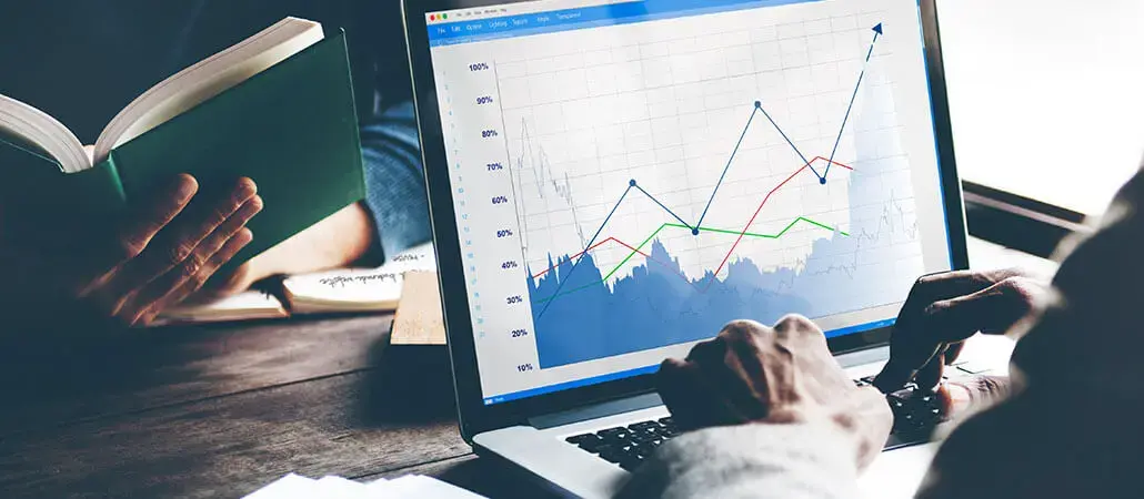 Persona utilizando una laptop con gráficos financieros de líneas y porcentajes en pantalla, mientras otra sostiene un libro abierto en segundo plano.