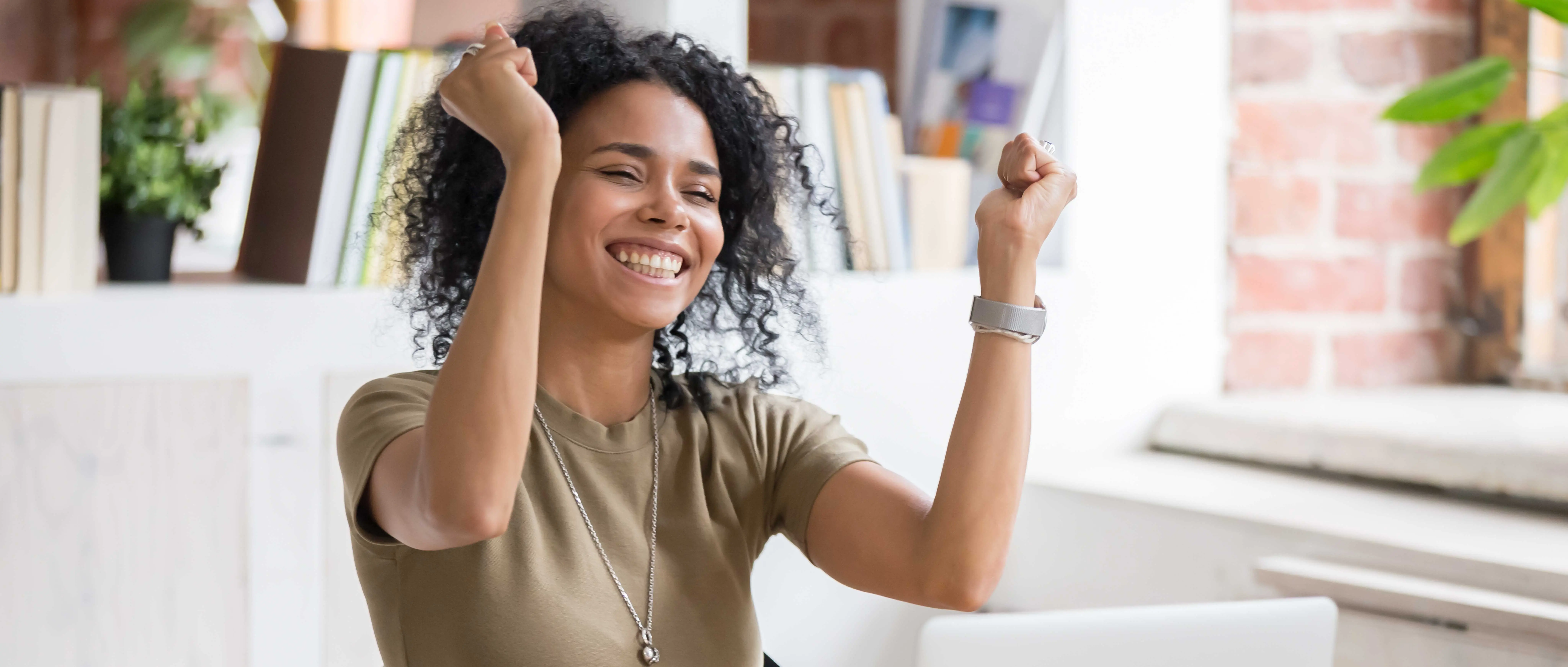 Mujer de cabello rizado levantando los brazos con entusiasmo frente a una computadora portátil en un espacio de oficina.
