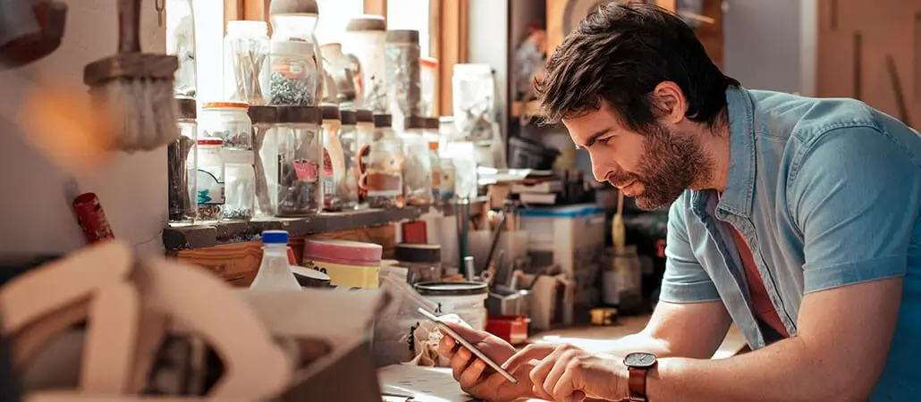 Hombre en taller observando su teléfono móvil mientras trabaja en una mesa con frascos y herramientas.