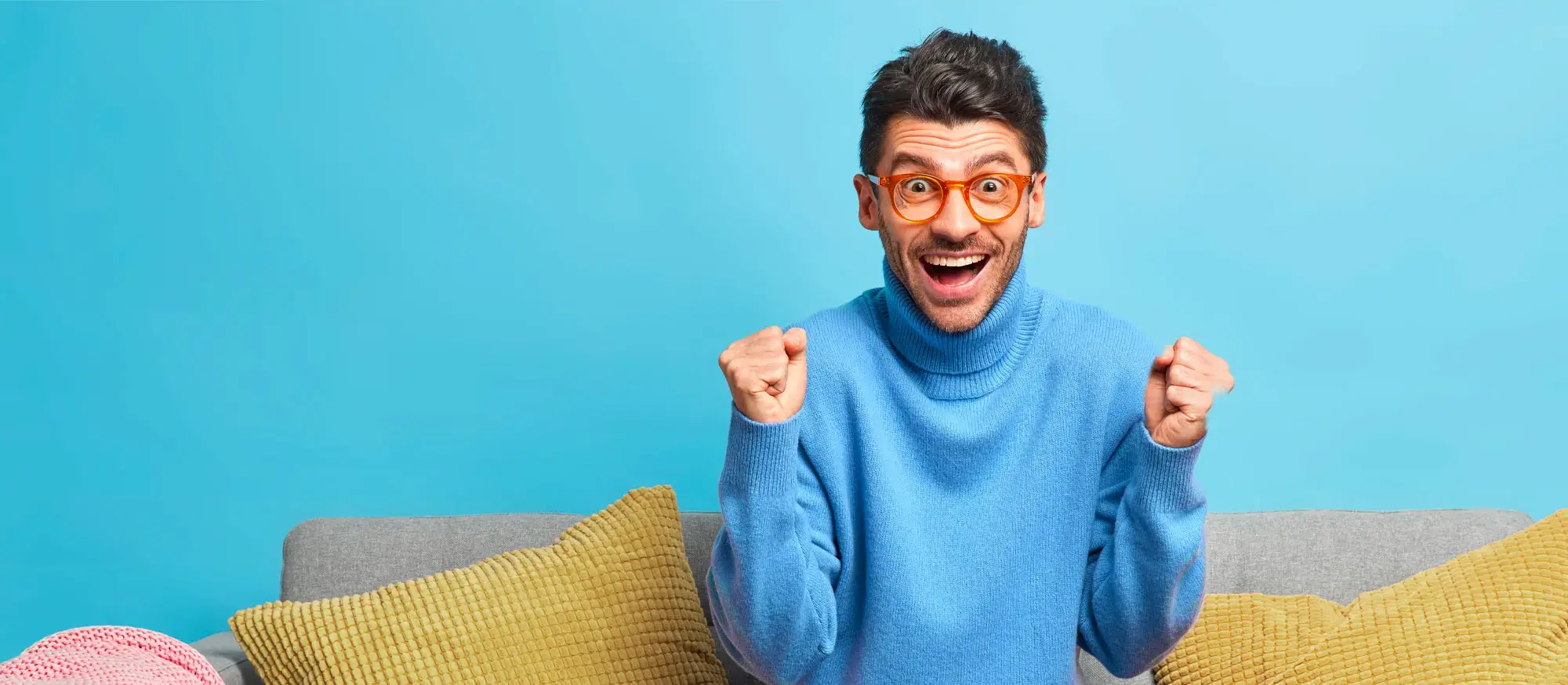 Hombre emocionado con gafas naranjas y suéter azul, sentado en un sofá gris con cojines, celebrando con los puños cerrados frente a un fondo azul.