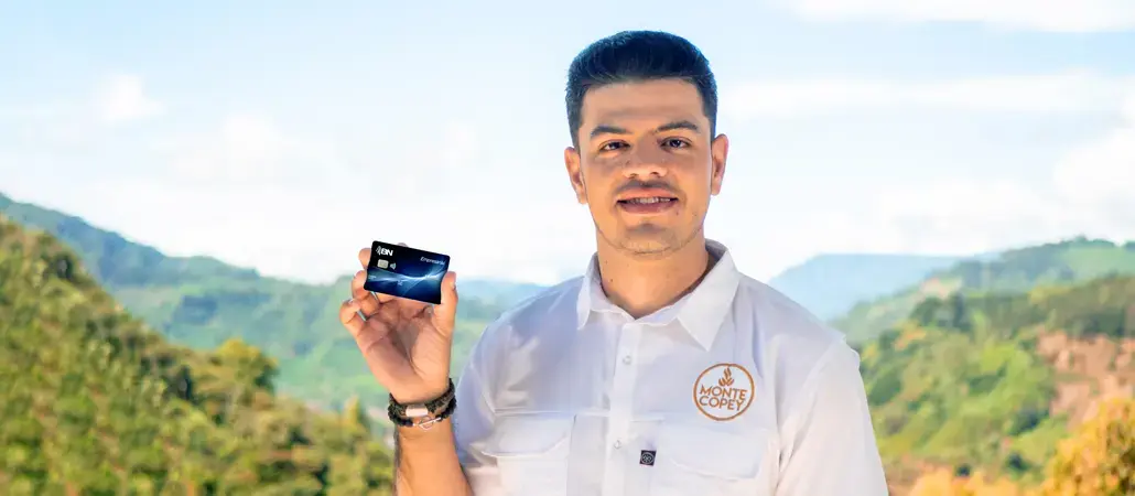 Hombre sonriente con camisa blanca de Monte Copey mostrando una tarjeta de crédito BN frente a un paisaje natural.