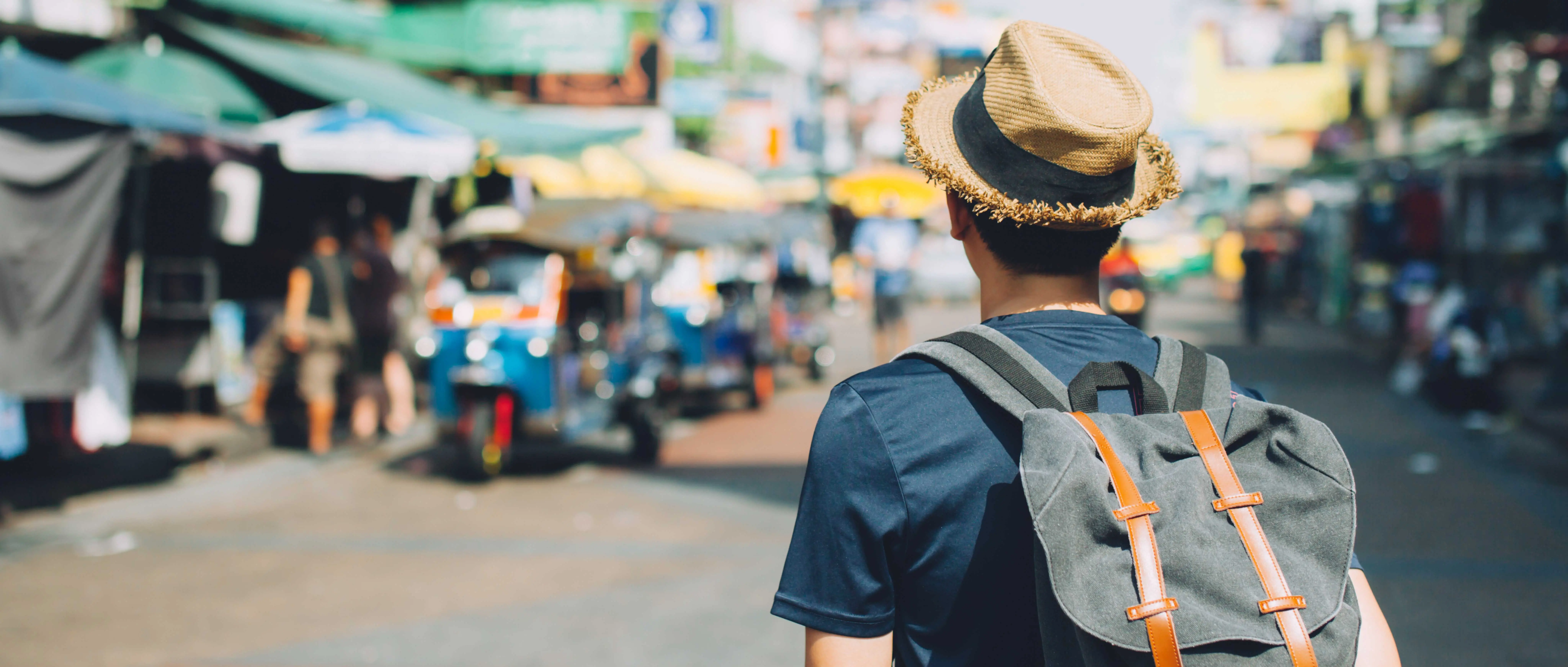 Persona con sombrero de paja y mochila negra caminando por una calle urbana con puestos y vehículos desenfocados al fondo.