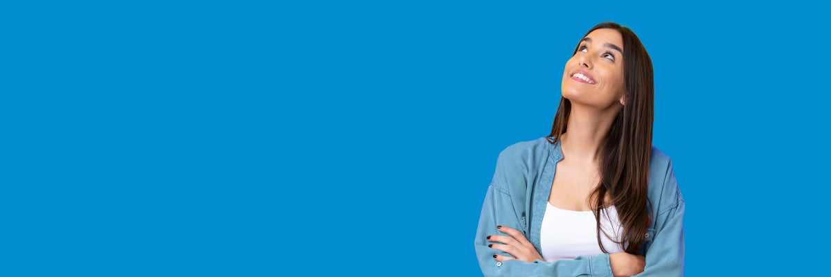 Mujer joven con camisa de mezclilla cruzada de brazos, mirando hacia arriba con expresión de esperanza sobre fondo azul sólido.