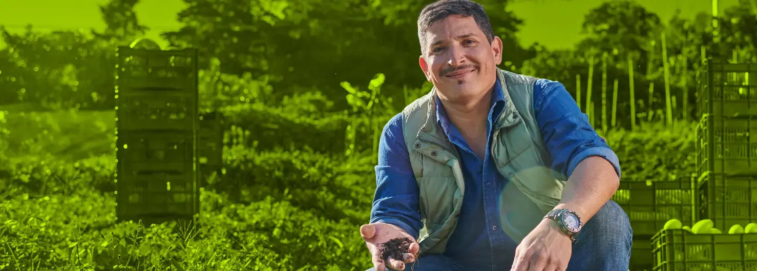 Hombre agricultor sonriente con camisa azul y chaleco verde, mostrando tierra en la mano, en un campo de cultivo con cajas plásticas apiladas al fondo.