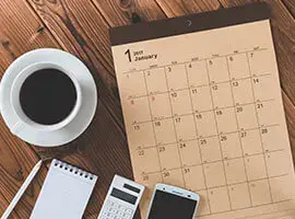 Calendario de enero sobre una mesa de madera, acompañado de una taza de café, libreta, calculadora y celular.